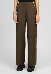 Pantalons larges en tissu vert olive foncé, dotés de boutons décoratifs sur les côtés et d'une texture lisse, associés à des chaussures à plateforme noires.