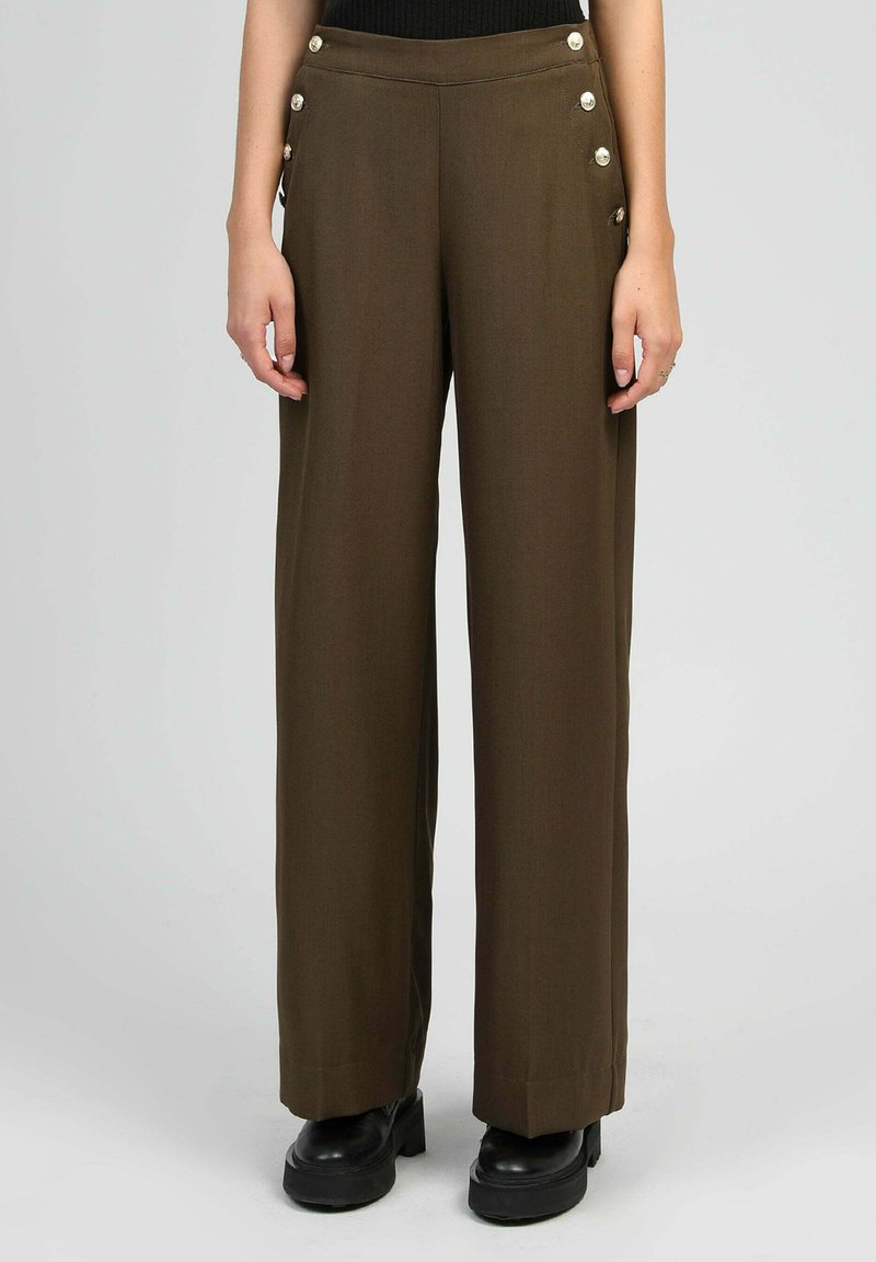 Pantalons larges en tissu vert olive foncé, dotés de boutons décoratifs sur les côtés et d'une texture lisse, associés à des chaussures à plateforme noires.