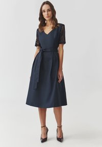 Marineblaues Midi-Kleid mit kurzen Spitzenärmeln, V-Ausschnitt, Taillengürtel und A-linienförmigem Rock. Glänzende schwarze Highheels ergänzen das Outfit.