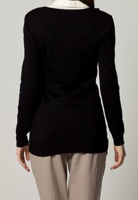 Black long-sleeve sweater with a scoop neckline, fitted shape, and ribbed hem, worn over light-colored trousers. Smooth texture.