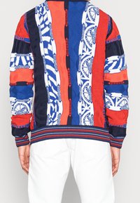 Gebreide trui met een patchworkontwerp in de kleuren rood, blauw en wit. De textuurpatronen bevatten geometrische en abstracte motieven.