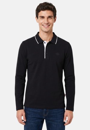 Homme souriant portant un polo noir à manches longues avec une bordure blanche au col et un jean bleu, debout devant un fond uni.