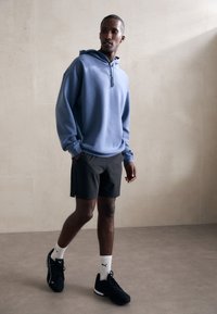 Blå hættetrøje med lynlås foran, parret med sorte shorts. Båret med sorte sko og hvide sokker med et logo. Glat stoftekstur.