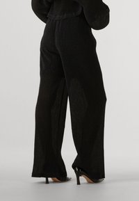 Zwarte wijde broek van textuur ribstof, met een hoge taille en zijsplitten, gecombineerd met zwarte hoge hakken.