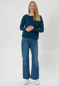 Türkiser, gerippter Langarmpullover mit quadratischem Ausschnitt, kombiniert mit hellblauen Schlagjeans und weißen Sneakers. Insgesamt glatte Textur.