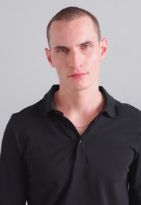 Cămașă polo neagră cu guler și mâneci scurte, cu un croi slim și textură netedă. Modelul are părul scurt și deschis la culoare, cu un fundal neutru.
