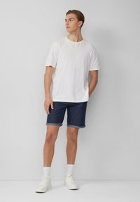 Weißes Baumwoll-T-Shirt kombiniert mit dunkelblauen Jeansshorts, die aufgerollte Bündchen haben. Model trägt weiße Sneakers und Knöchelsocken.