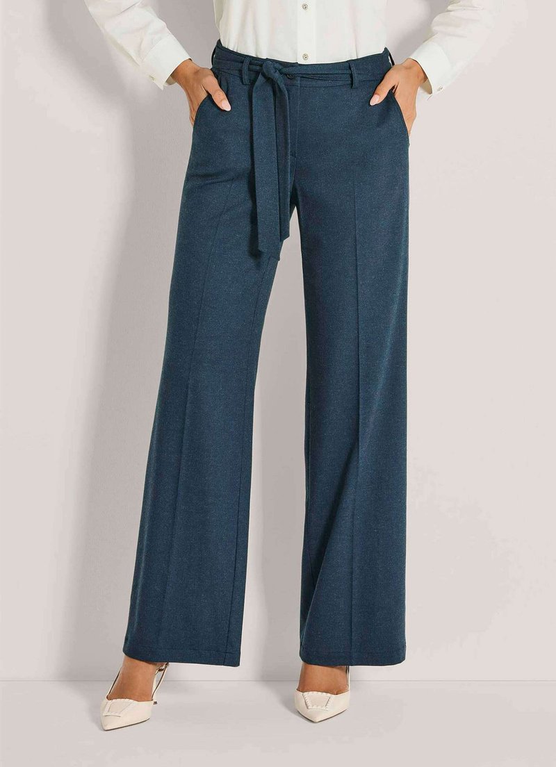 Vrouw in hoge taille donkergroene wijde broek met bijpassende riem, witte blouse en witte puntige hakken.