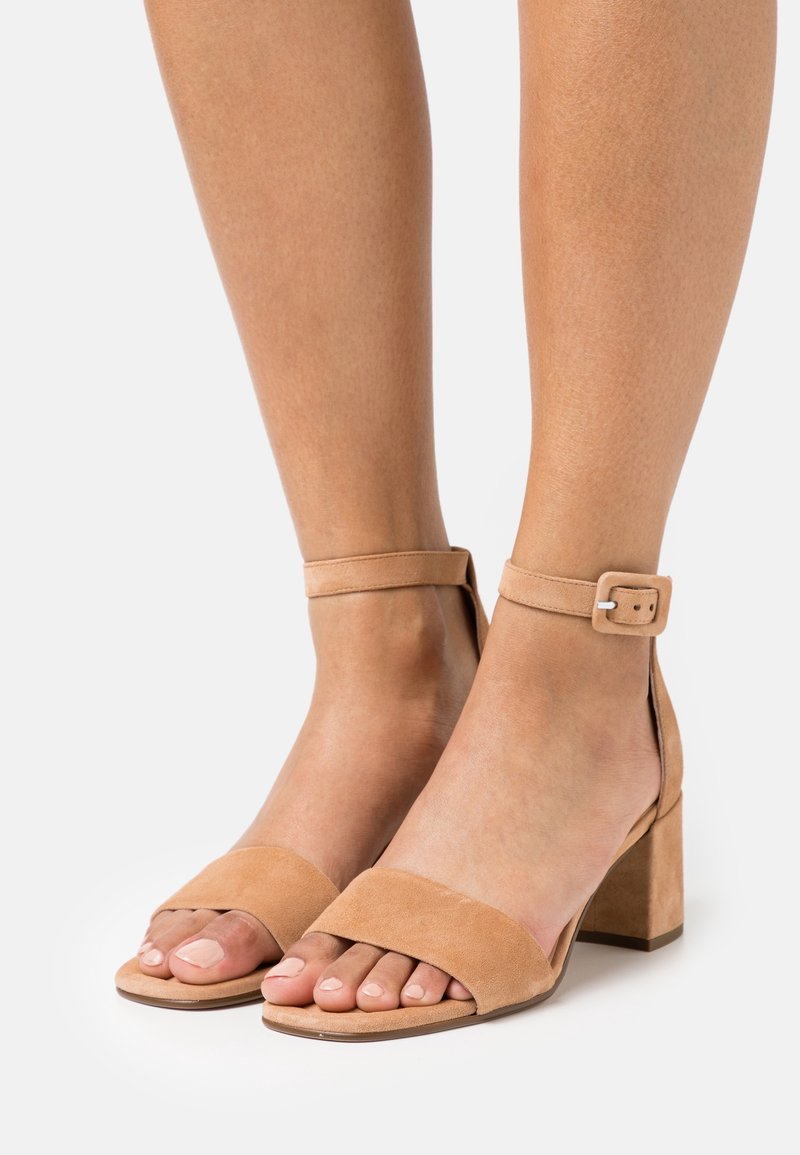 Pieds portant des sandales à talon bloc en suède beige avec brides à la cheville et bouts carrés sur fond blanc uni.