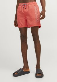 Bañador de coral con cintura elástica y cordón. Detalle gráfico en blanco "JACK&" en la pierna izquierda. Combinado con sandalias deslizantes negras.
