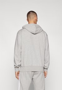 Hoodie cinza com capuz ajustável, punhos e barra canelados. Textura suave, corte descontraído. Usado com calças de moletom cinza combinando, apresentando riscas laterais.