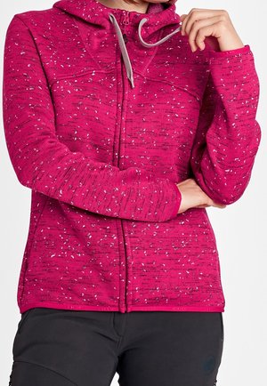 Mujer con una sudadera con capucha rosa moteada y cremallera, con cordones grises y pantalones negros, con los brazos cruzados y la mano tocando la barbilla.