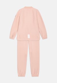 Ružový pletený teplákový komplet pozostávajúci z mikiny na zips s vzormi v tvare V a zodpovedajúcich nohavíc s elastickými manžetami a logom na zadnej strane.
