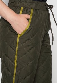 Gequilte groene broek met gele rand bij de zakken, voorzien van een elastische tailleband en een verstelbaar trekkoord, met een gladde textuur.