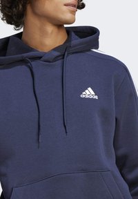 Laivastonsininen huppari kengurutaskulla, jossa on kiristysnauhallinen huppu ja valkoinen Adidas-logo rinnassa. Pehmeä kangas ja pelkistetty muotoilu.
