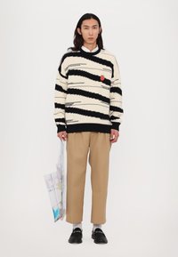Tröja med svarta och krämfärgade zigzag-ränder; har en röd patch, bärs med beige byxor och svarta loafers, håller en genomskinlig väska.