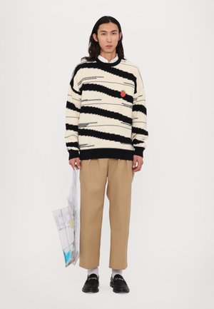 Pullover mit schwarzen und cremefarbenen Zickzackstreifen; verfügt über einen roten Patch, getragen mit beigen Hosen und schwarzen Loafers, hält eine transparente Tasche.