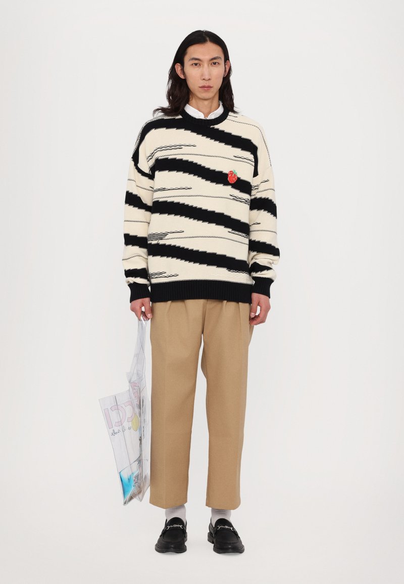 Tröja med svarta och krämfärgade zigzag-ränder; har en röd patch, bärs med beige byxor och svarta loafers, håller en genomskinlig väska.