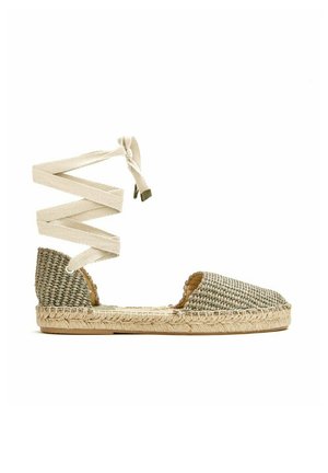 Sandalia plana estilo espadrille con parte superior de tela tejida y cordones para atar en el tobillo en beige con suela de yute natural sobre fondo blanco.