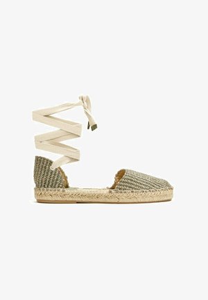 Sandalia plana estilo espadrille con parte superior de tela tejida y cordones para atar en el tobillo en beige con suela de yute natural sobre fondo blanco.