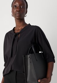 Zwarte blouse met een strikdetail bij de hals en een gestructureerde stof, gecombineerd met een gestructureerde zwarte handtas met een subtiel patroon aan de buitenkant en korte handvatten.