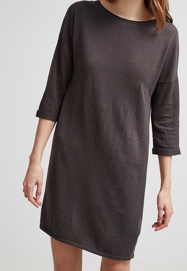 Femme portant une robe simple gris foncé, longueur genou, avec des manches retroussées au trois-quarts, sur un fond clair uni.