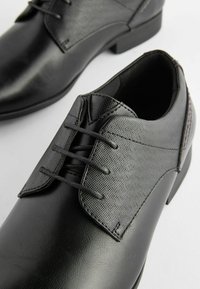 Zapatos de vestir negros con puntera de cuero suave y parte superior de tela texturizada. Cuatro ojetes para los cordones, con un tacón de perfil bajo.