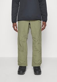 Pantalons de neige vert olive avec une coupe ample, fabriqués en matériau synthétique, dotés de poches zippées et de coutures renforcées. Des bottes de neige noires sont portées.