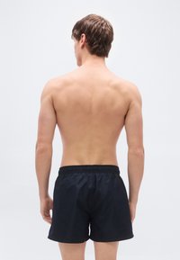  Μαύρα μαγιό σορτς με ελαστική μέση, λεία υφή και χωρίς ορατά σχέδια, εμφανιζόμενα από πίσω. Ορατός φυσικός τόνος δέρματος.