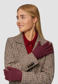 Gants tricotés bordeaux avec une texture lisse, présentés sur un modèle portant un blazer en tweed à motifs et une chemise bourgogne en dessous.