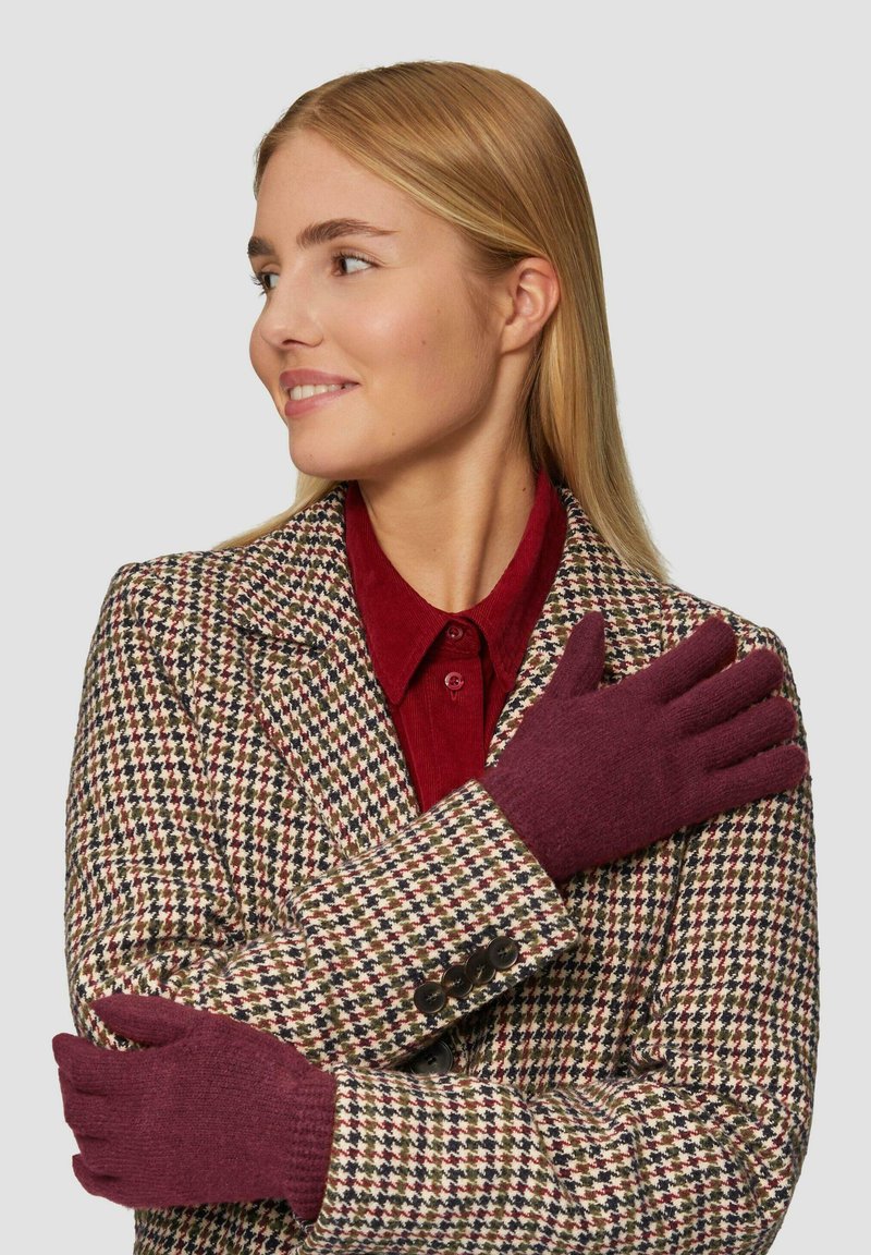 Gants tricotés bordeaux avec une texture lisse, présentés sur un modèle portant un blazer en tweed à motifs et une chemise bourgogne en dessous.