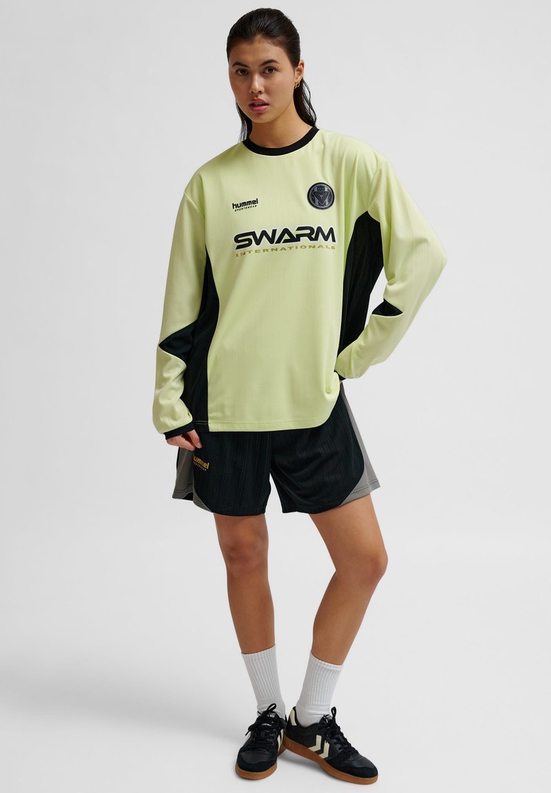 Lichtgroene sporttop met lange mouwen en zwarte accenten, met het logo "SWARM", gecombineerd met zwarte shorts en witte sokken. Zwarte sneakers maken de look af.