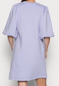 Femme portant une robe lilas courte et ample avec des manches bouffantes à mi-avant, vue de dos sur un fond uni.