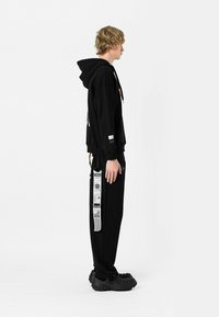 Sudadera negra con bolsillo tipo canguro y cordón, combinada con pantalones jogger negros que tienen acentos gráficos blancos a lo largo de los lados. Zapatos negros.