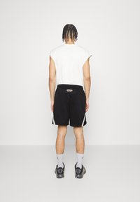 Schwarze Sportshorts mit einem weißen Streifen, kombiniert mit einem weißen ärmellosen Shirt. Getragen mit grauen Socken und schwarzen Sportschuhen.