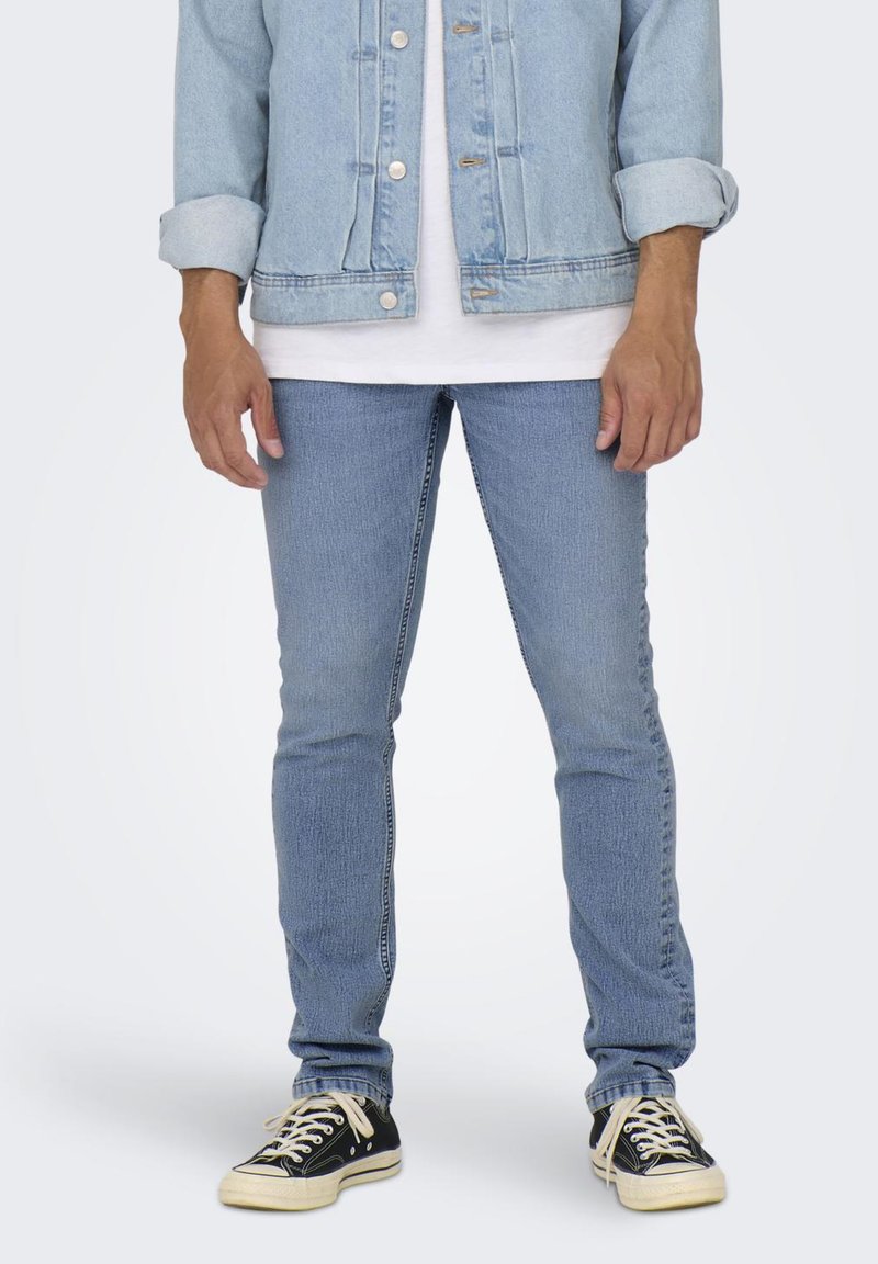 Lys blå denimjakke og tettsittende lys blå jeans, brukt sammen med en hvit t-skjorte og svarte joggesko. Detaljer inkluderer metallknapper og sømmer.