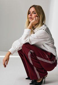 Camicia bianca con dettagli di bottoni sulle spalle, abbinata a pantaloni bordeaux con righe bianche e tacchi neri. Modella posata su uno sfondo bianco.