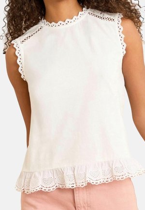 Biały top bez rękawów z falbankowym obszyciem z ażurowej koronki na dekolcie, ramionach i falbaniastym dole, noszony z bladoróżowymi spodniami.