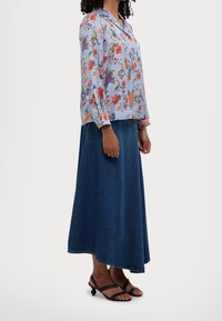 Blouse à motif floral en bleu clair, avec des manches longues et des poignets boutonnés, accompagnée d'une jupe maxi tiered en denim foncé et de sandales noires.
