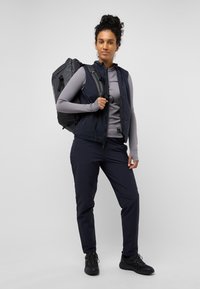 Gilet blu sopra una maglietta grigia a maniche lunghe, abbinato a pantaloni e scarpe neri. Zaino nero con spallacci regolabili sulla schiena del modello.