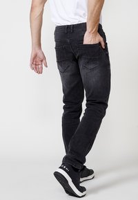 Jeans de denim negros ajustados con un ligero desgaste, con bolsillos traseros y una etiqueta en la cinturilla, combinados con zapatillas negras.