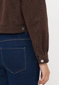 Bruine denimjas met luipaardprint en knoopmanchetten, gecombineerd met donkerblauwe jeans met een gebogen stiksels in de achterzak.