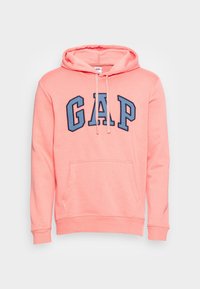 Sudadera rosa con letras azules "GAP" en el pecho, bolsillo canguro en la parte frontal y cordones en la capucha, presentada sobre un fondo blanco.