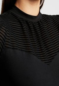 Schwarzes Oberteil mit hohem Kragen, einem transparenten oberen Bereich mit schwarzen Streifen und einem undurchsichtigen schwarzen unteren Bereich mit leicht entspanntem Sitz.