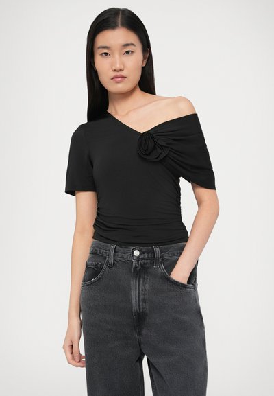 Μαύρο ασύμμετρο τοπ με κοντά μανίκια, με λεπτομέρεια τριαντάφυλλου στον ώμο. Φοριέται με ψηλόμεσο σκούρο γκρι τζιν. Λεία υφή υφάσματος.