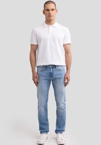 Mees valges polo-särgis, helesinistes teksapükstes ja valgetes tossudes, seisab ühevärvilise heledalt halli tausta ees.