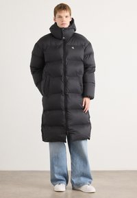 Sort pufferjakke med høj krave, der har quiltede sektioner, en frontlynlås og sidelommer. Parret med lyseblå wide-leg jeans.