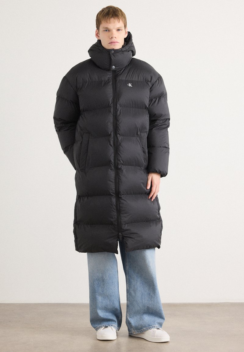 Sort pufferjakke med høj krave, der har quiltede sektioner, en frontlynlås og sidelommer. Parret med lyseblå wide-leg jeans.