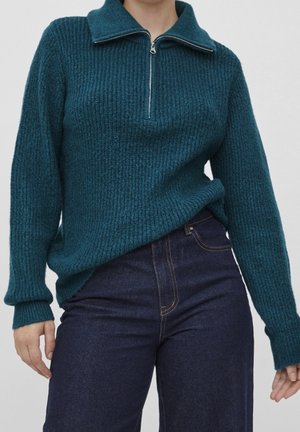 Teal gebreide trui met een hoge kraag en een halve ritssluiting. Geribbelde textuur en lange mouwen, gecombineerd met een donkere spijkerbroek.
