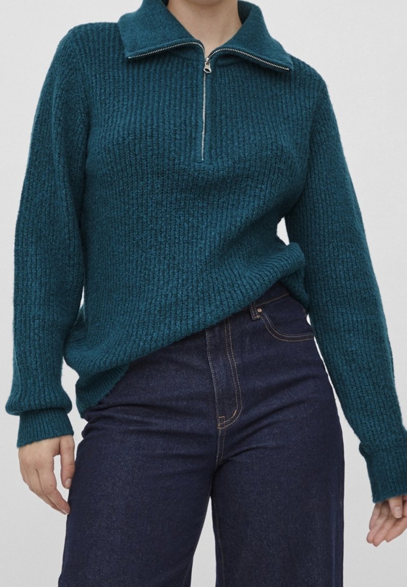 Pull en maille bleu canard avec un col montant et une fermeture éclair mi-longue. Texture côtelée et manches longues, associé à un jean en denim foncé.